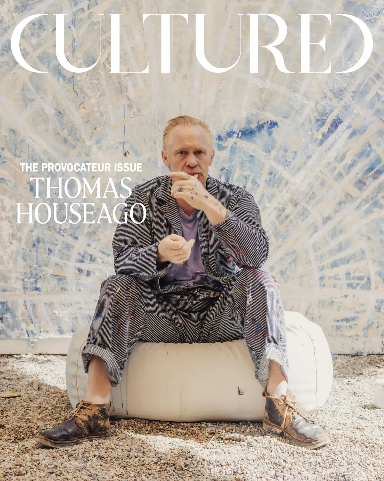 Portrait of British sculptor Thomas Houseago seated on a white sculptural cushion in his paint-splattered studio, with a monumental abstract blue-and-white canvas behind him for the cover of Cultured magazine's 'The Provocateur Issue.'