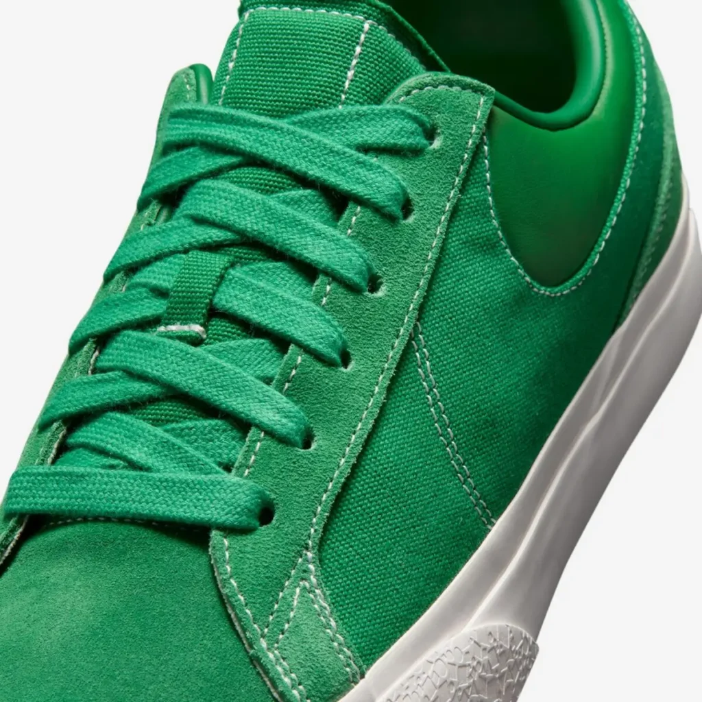 Close-up of the forefoot showcasing green suede overlays, textured canvas side panel, tonal flat laces, and contrast white stitch