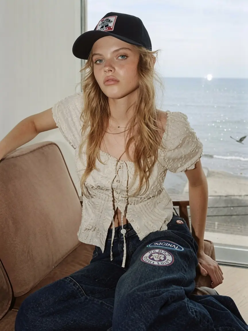 Blonde model wearing a black logo baseball cap, textured white puff-sleeve blouse, and wide-leg patched denim jeans while seated beside a window overlooking the ocean.