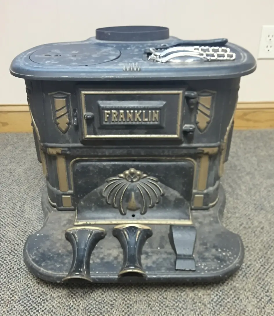 Vintage Franklin cast-iron parlor stove with black finish, brass trim, decorative front panels, and an embossed FRANKLIN nameplate, photographed indoors on gray carpet against a beige wall.