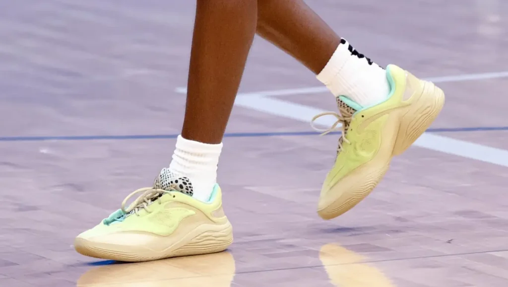 Close-up of a person wearing white socks and pastel-colored sneakers with light green, yellow, and beige tones, walking on a polished indoor basketball court.