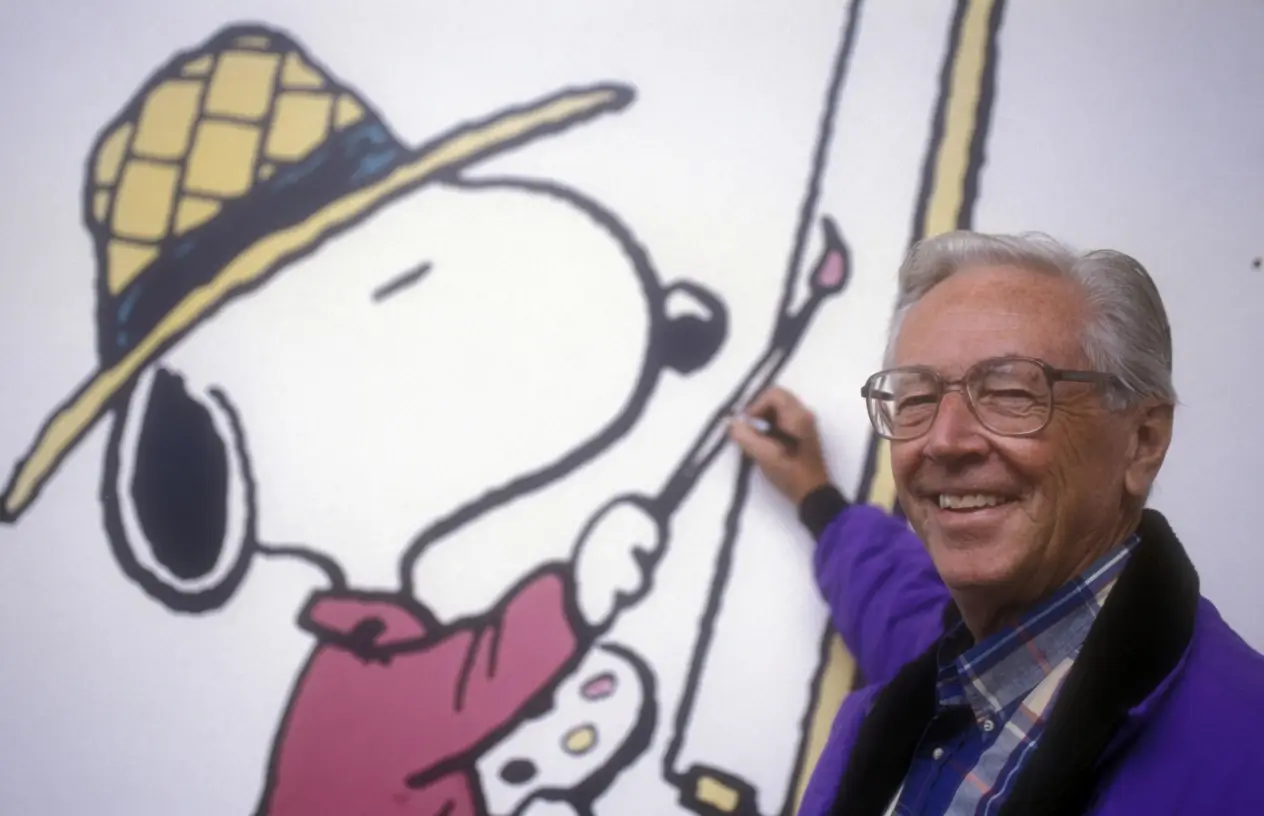 Charles M. Schulz smiles beside a large mural of Snoopy painting while holding a brush, highlighting the beloved Peanuts creator alongside his iconic character.