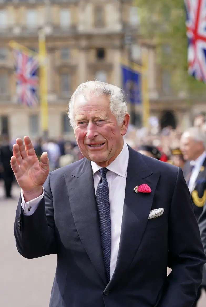 A formally dressed man waves to onlookers during a ceremonial event, wearing a dark navy suit, white dress shirt, patterned tie, white pocket square, and a bright pink boutonnière. Union Jack flags line the street behind him as historic buildings and a gathered crowd create a festive backdrop.