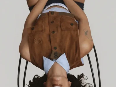 Upside down studio portrait of beabadoobee seated on a black metal chair, wearing a light blue collared shirt beneath a distressed brown suede vest embossed with a tonal logo. The model has curly dark hair, glossy lips, visible arm tattoos, black shorts, white waistband detailing, and patterned tights with circular brown and cream motifs, posed against a clean light gray background