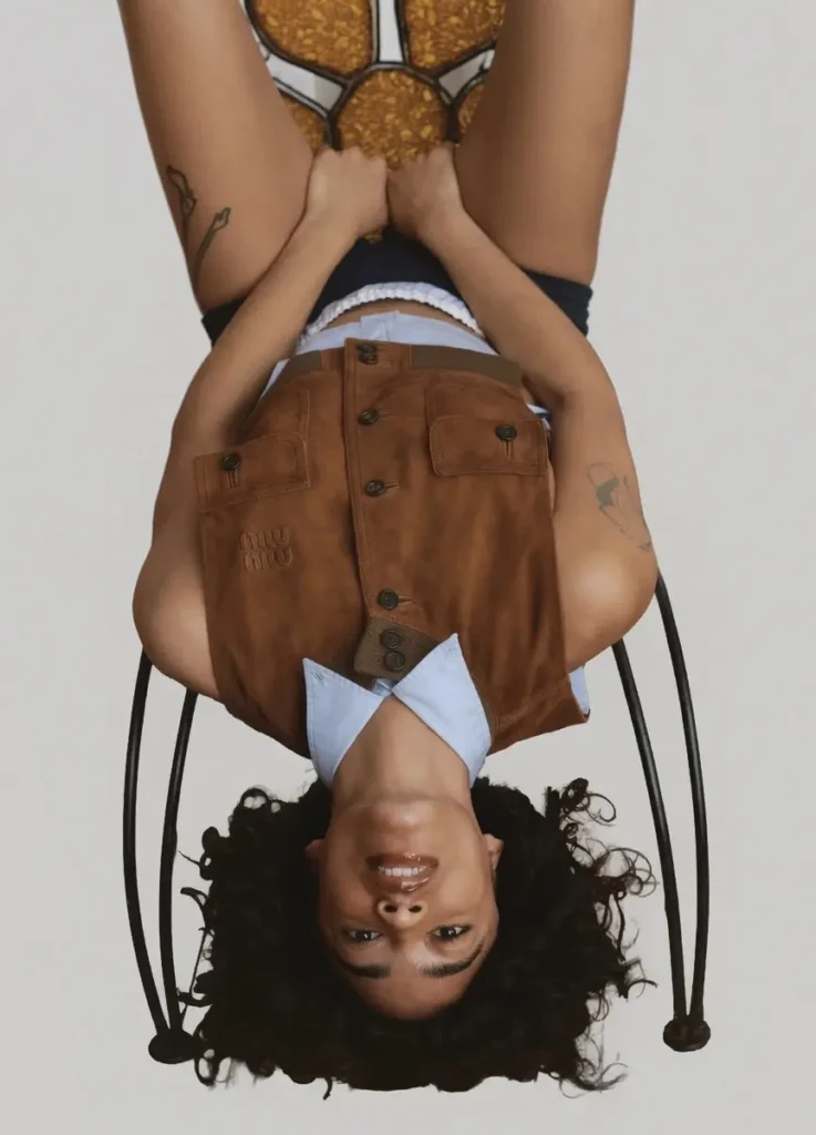 Upside down studio portrait of beabadoobee seated on a black metal chair, wearing a light blue collared shirt beneath a distressed brown suede vest embossed with a tonal logo. The model has curly dark hair, glossy lips, visible arm tattoos, black shorts, white waistband detailing, and patterned tights with circular brown and cream motifs, posed against a clean light gray background