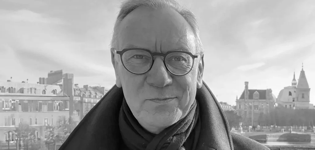 Black-and-white portrait of Hugh Devlin wearing round glasses, a dark overcoat, and a scarf, standing outdoors with historic European architecture, a bridge, and a waterfront softly blurred behind him under a bright, cloud-filled sky