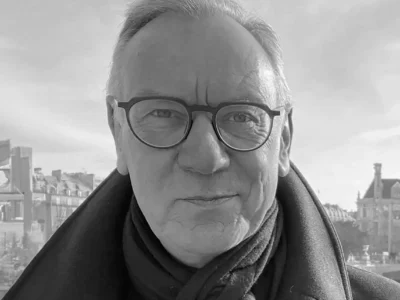 Black-and-white portrait of Hugh Devlin wearing round glasses, a dark overcoat, and a scarf, standing outdoors with historic European architecture, a bridge, and a waterfront softly blurred behind him under a bright, cloud-filled sky