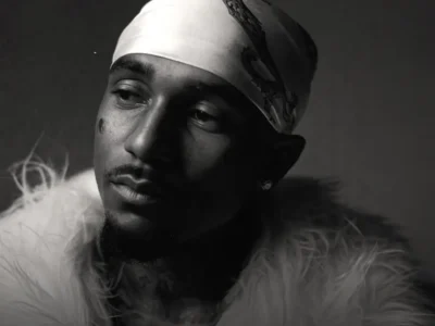 A monochrome portrait of a Loe Shimmy wearing a white printed durag, a fluffy faux-fur jacket, stud earrings, and a chain necklace. He looks off to the side with a contemplative expression against a dark studio background, while dramatic side lighting emphasizes his facial features and the high-contrast black-and-white