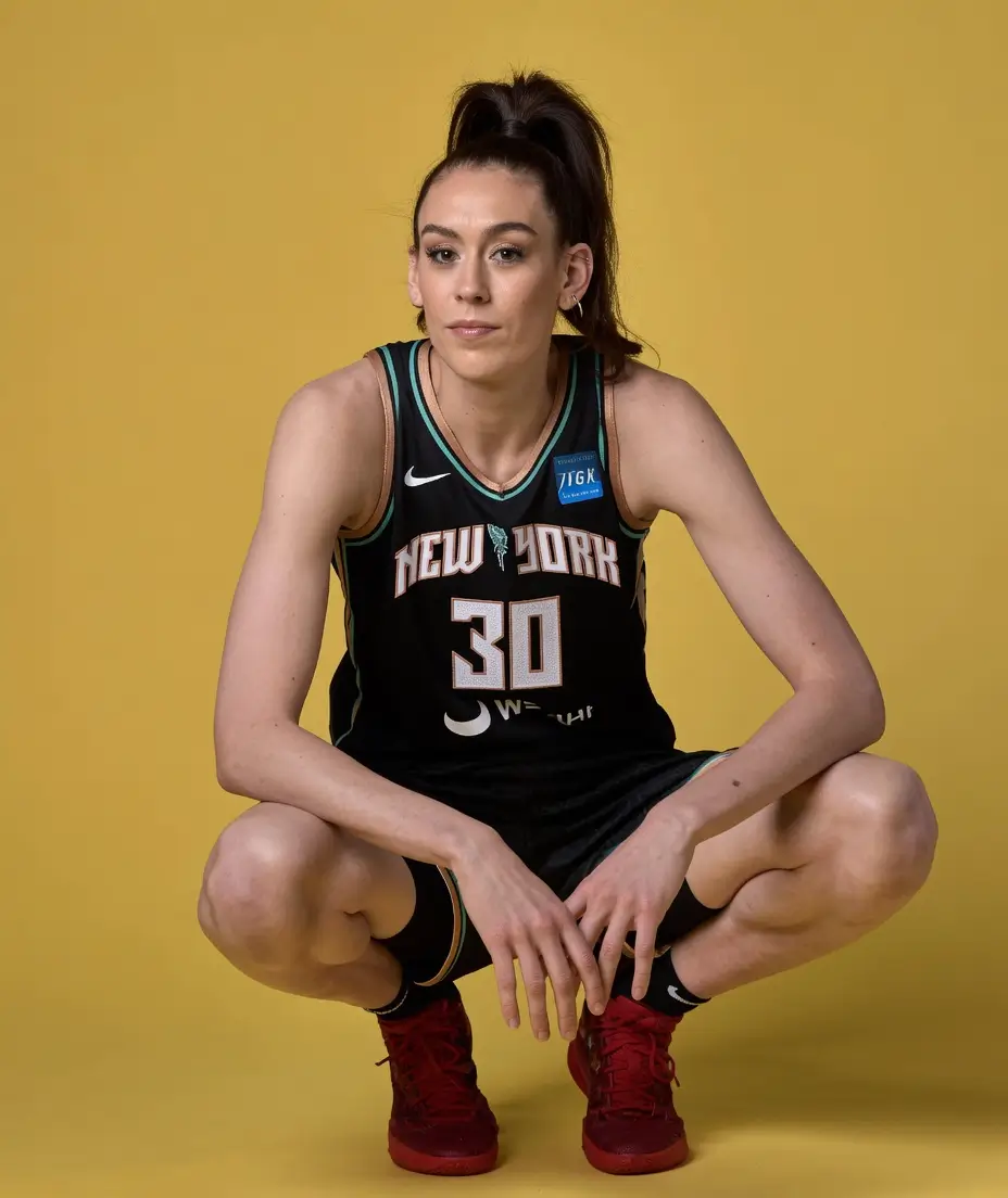 Professional studio portrait of a New York Liberty basketball player crouching against a mustard-yellow backdrop, wearing the team's black road uniform with number 30, black socks, and bright red basketball shoes. The athlete has a high ponytail and looks directly toward the camera in a composed media day pose.