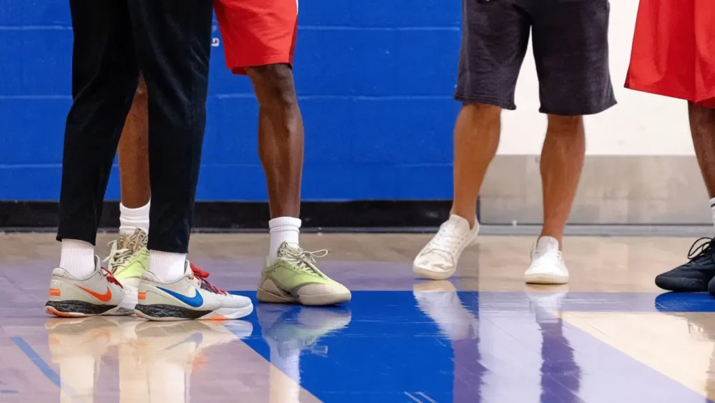 Three people stand on an indoor basketball court. Only their legs and shoes are visible, each person wearing different athletic sneakers and shorts. The floor is polished, and the wall behind them is blue.