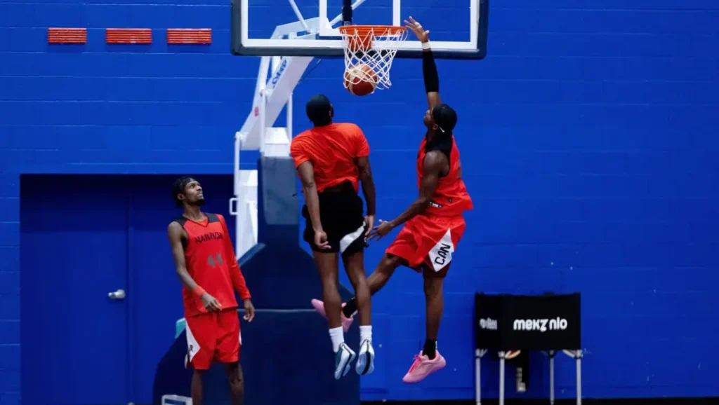 Three basketball players in red uniforms play in a gym. One jumps to block a shot as another attempts a layup, with the ball just touching the rim. The third player stands nearby, watching the action. Blue wall and hoop in the background.