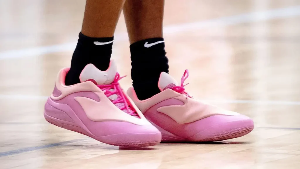 Close-up of a person wearing pink and light pink sneakers with black Nike socks, standing on a wooden indoor court.
