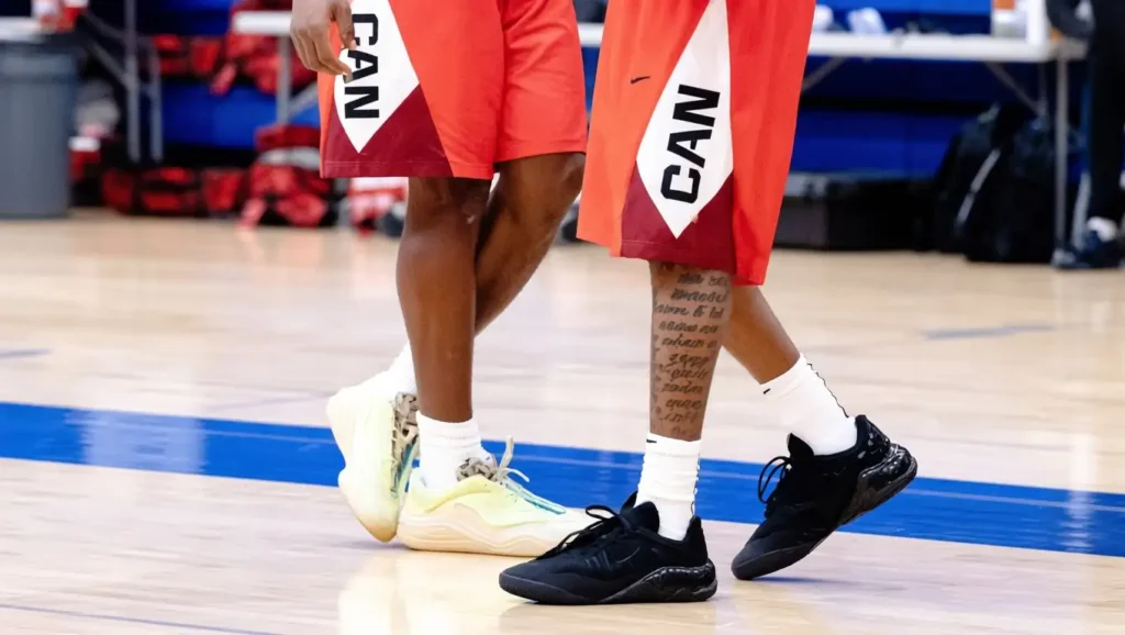 Two basketball players wearing red shorts labeled CAN stand on a gym court. One has a leg tattoo and black shoes; the other has white shoes with blue accents. Equipment bags are visible in the background.