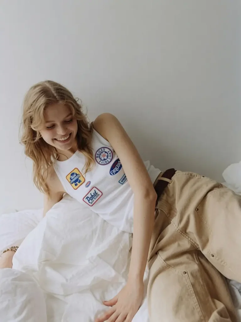 Smiling blonde model reclining on white bedding wearing a ribbed white patch-covered tank top and relaxed khaki pants in a minimalist bedroom