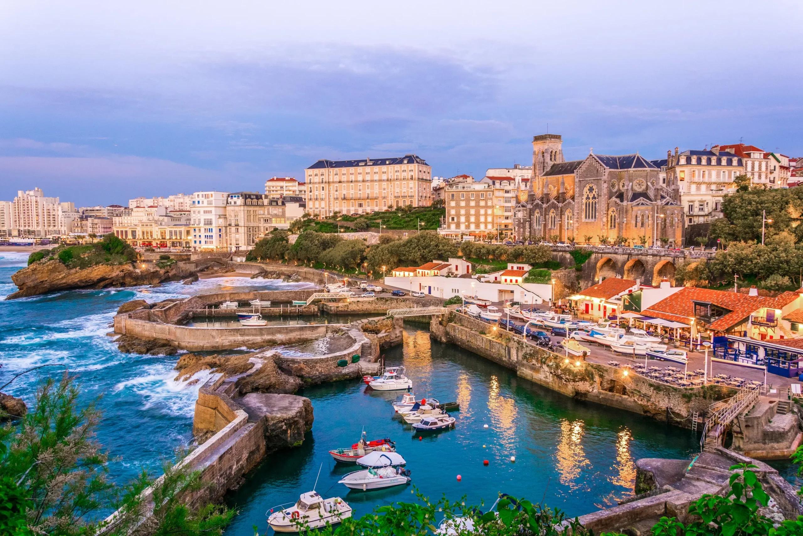 Aerial dusk view of the Port des Pêcheurs in Biarritz, France, showing small fishing and pleasure boats moored in a rocky natural harbor, with Sainte-Eugénie church and the town's coastal buildings illuminated against a twilight sky, and waves breaking against the cliffs beyond the harbor wall.