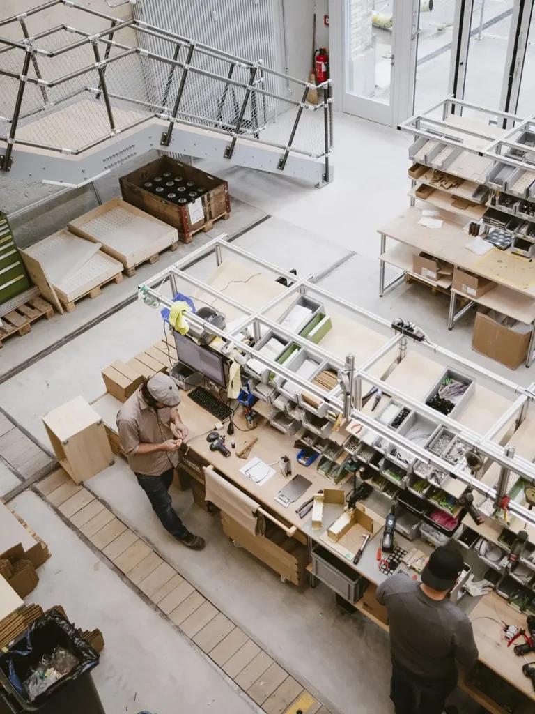 Overhead view of a modern espresso machine workshop where technicians assemble components at organized workstations inside a bright industrial production facility