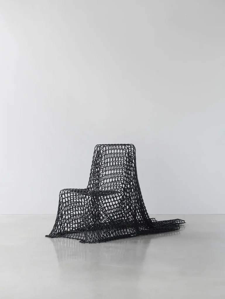 A sculpture chair wrapped in an open black lattice textile, its mesh draping over the seat and flowing onto the polished concrete floor against a minimalist white gallery backdrop