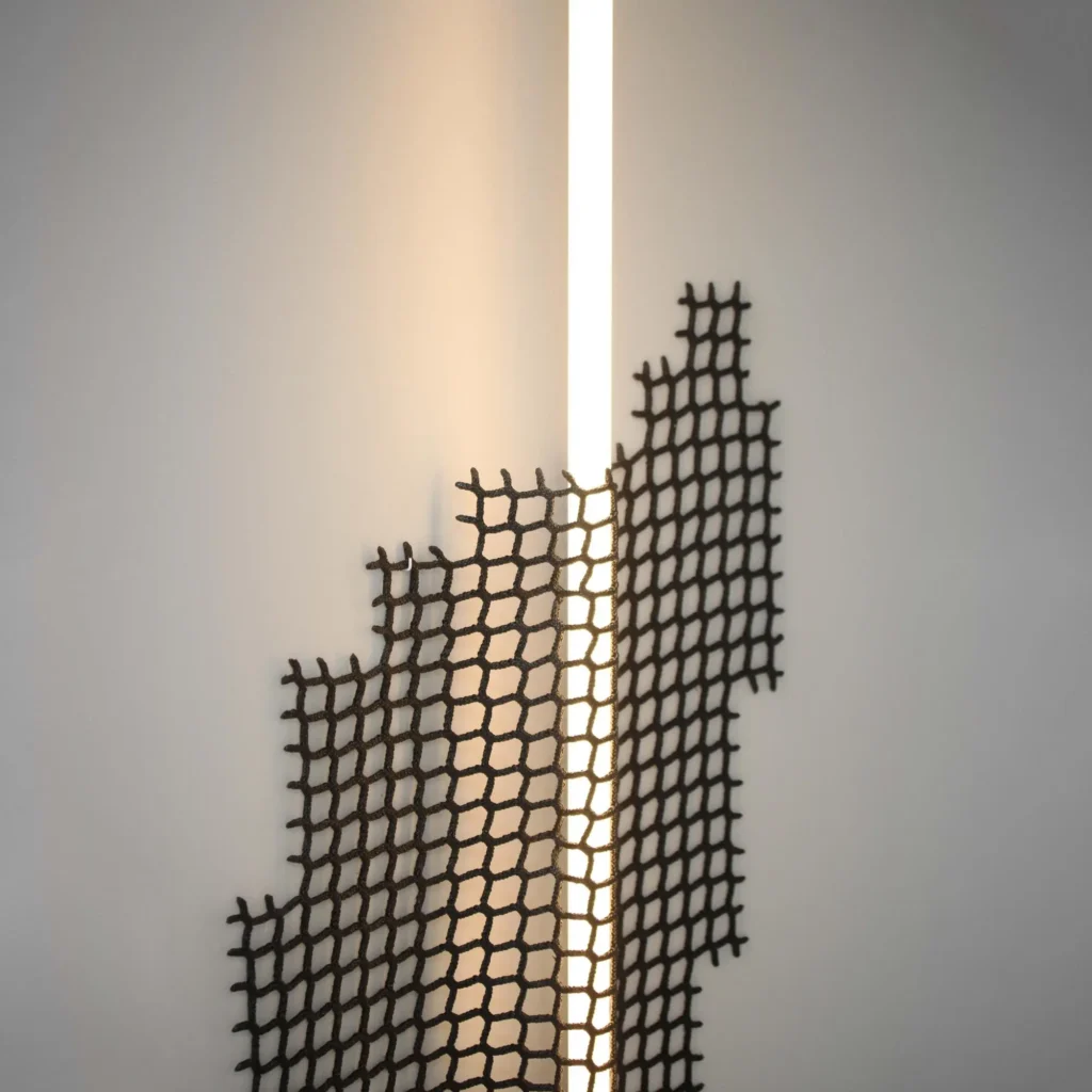 A vertical LED light strip framed by irregular black woven mesh panels, creating geometric shadows and soft warm illumination on a clean white wall