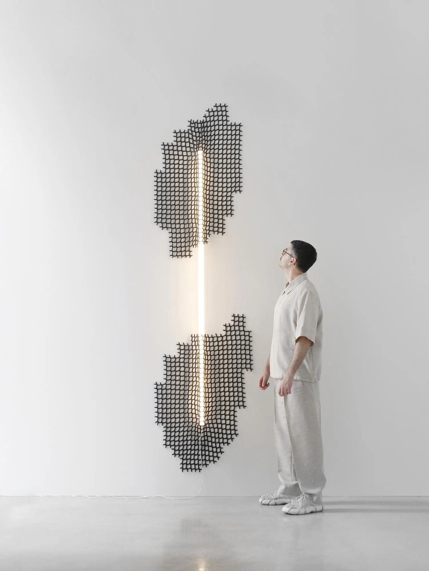 A visitor dressed in light neutral clothing stands beside a tall wall-mounted light sculpture composed of two warm white vertical LED tubes framed by irregular black woven mesh panels. Set against a minimalist white gallery wall, the installation emphasizes scale, texture, and the interplay of recycled netting with contemporary lighting design.