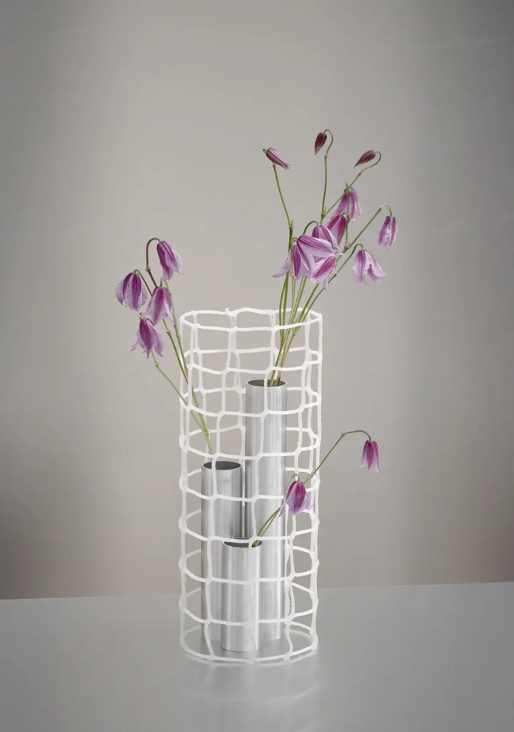 Contemporary floral vase: Purple bell-shaped flowers arranged in aluminum tube vases enclosed within a white woven cylindrical lattice vessel, displayed on a clean tabletop against a soft gray background
