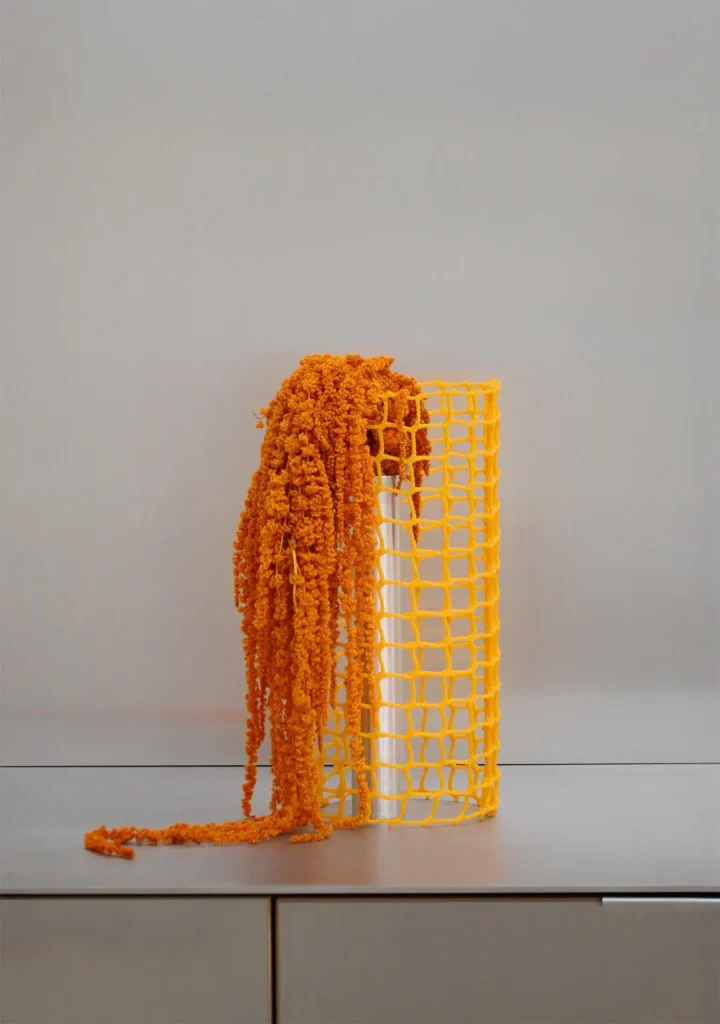 Yellow woven vessel with flowers: A bright yellow open-weave cylindrical vase holding cascading orange amaranthus blooms, with long trailing stems spilling over the edge onto a minimalist cabinet.