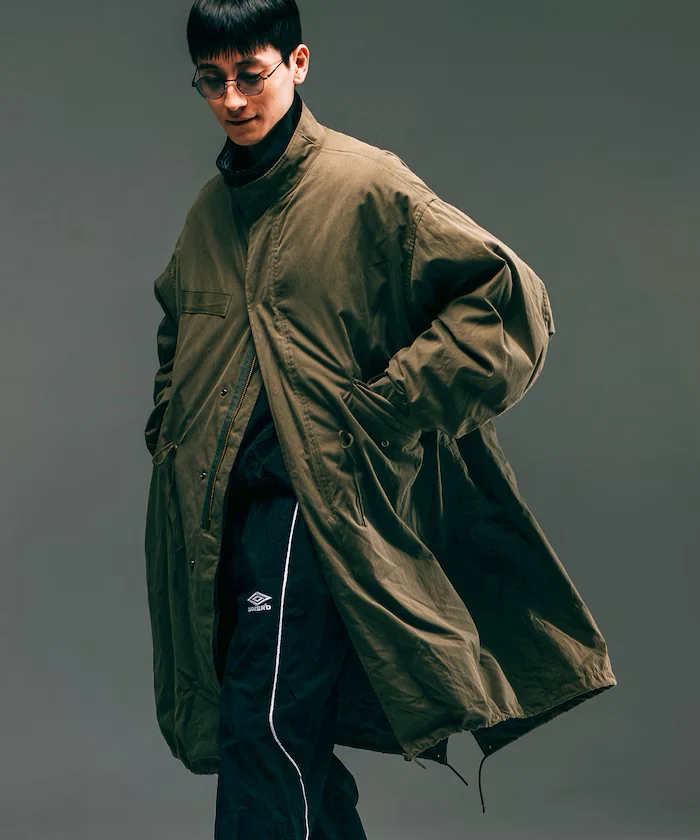 Model wearing an oversized olive military-inspired fishtail parka with a stand collar, concealed zip and snap closure, oversized flap pockets, adjustable drawcord waist, and flowing long back hem in a relaxed utilitarian silhouette.