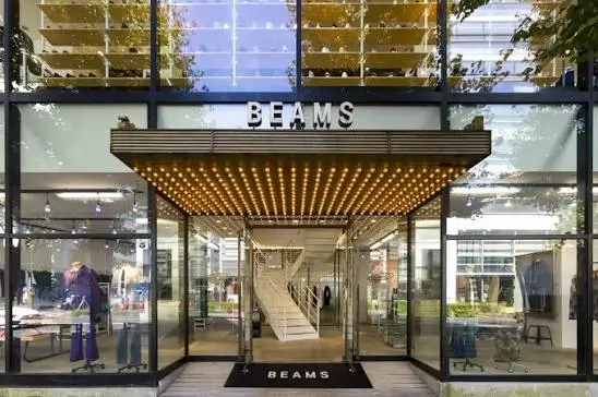 Exterior entrance of a BEAMS retail store featuring a glass façade, illuminated marquee canopy, and modern showroom visible through floor-to-ceiling windows.
