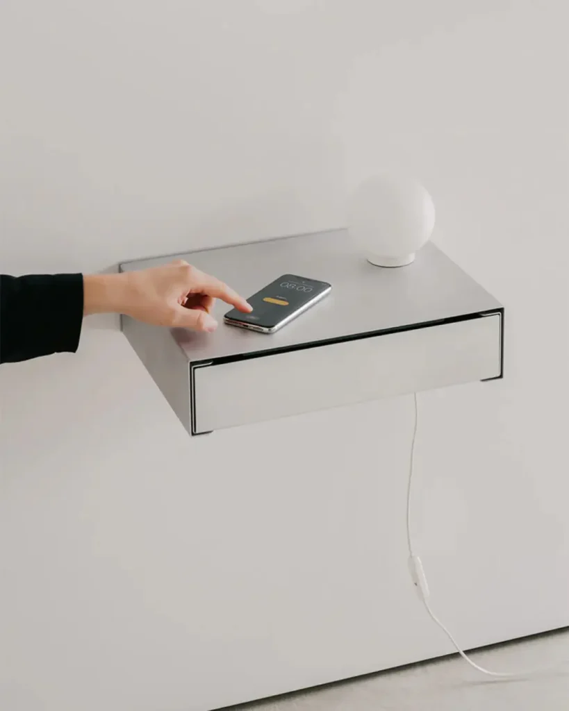 Silver wall-mounted floating drawer shelf holding a smartphone and small table lamp, with a hand reaching toward the phone
