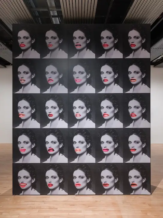 Installation view of a large grid artwork featuring repeated black-and-white female portraits, each altered with different oversized red lips, creating a pop art-inspired exploration of repetition, collage, and identity.