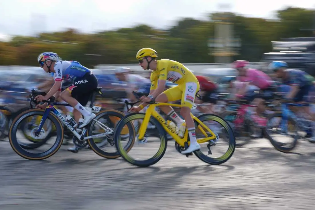 The Tour de France peloton rides through Paris during the final stage, with the race leader in the iconic yellow jersey positioned near the front alongside rival riders. Motion blur emphasizes the speed of the group as they race across cobblestone streets beneath clear summer skies.