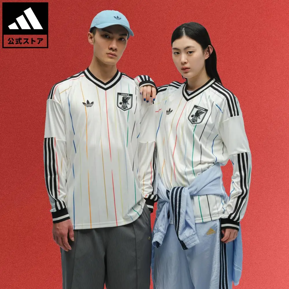 Two models wear the adidas Originals x Japan Football Association white away jersey, featuring multicolored vertical pinstripes, black three-stripe sleeves, the JFA crest, and Trefoil branding against a red studio backdrop.