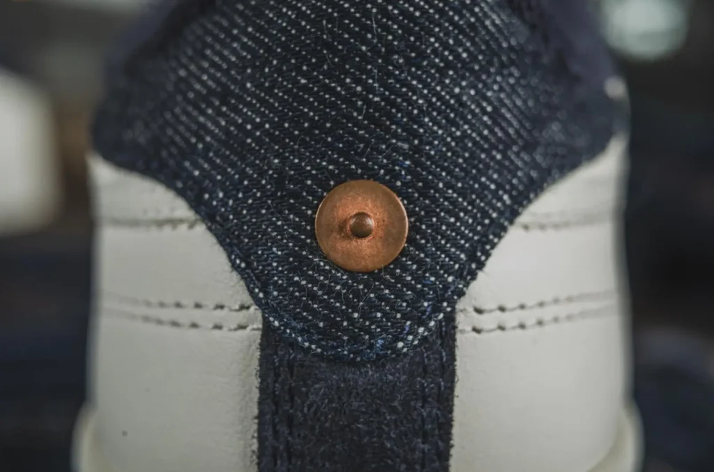 Close-up of the heel construction on the Reebok x TCB Jeans shoe, showing textured indigo selvedge denim, white leather heel panels, exposed stitching, and a copper rivet inspired by traditional denim hardware