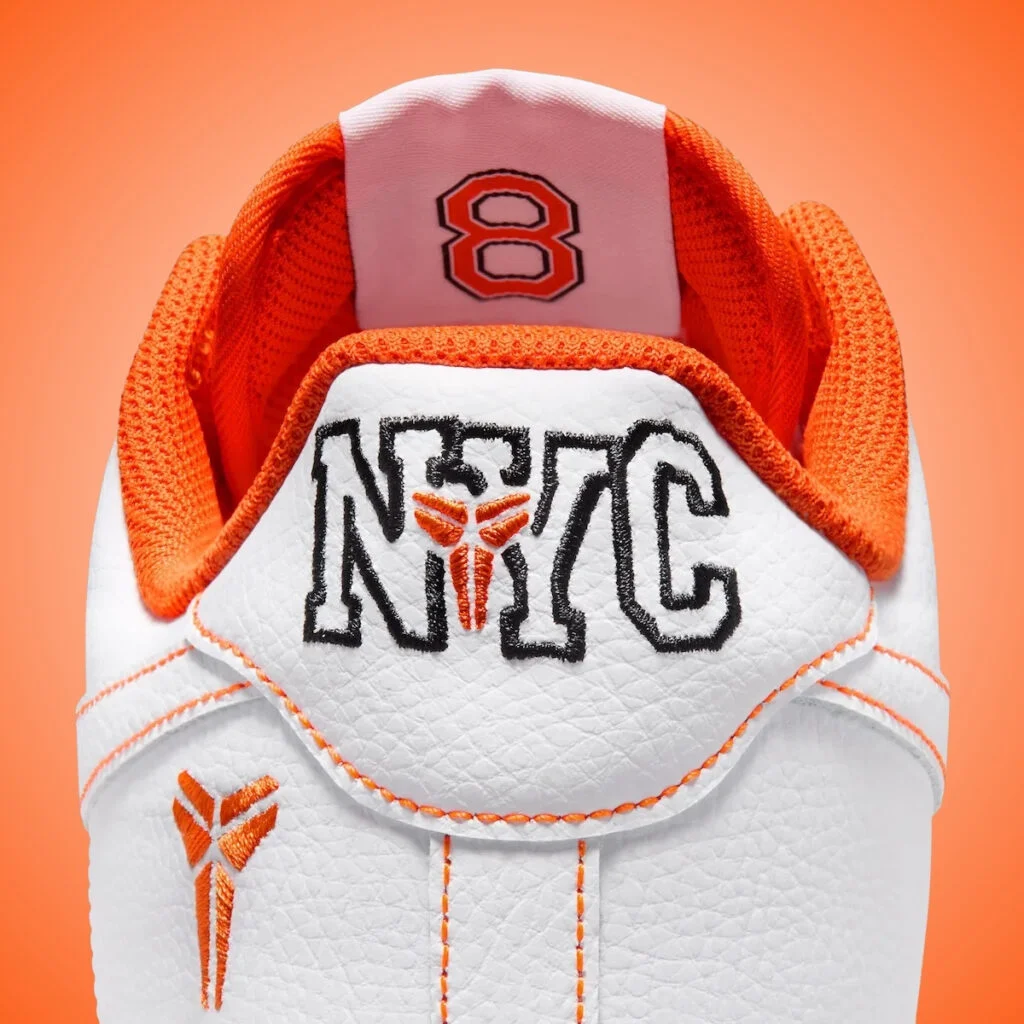 Close-up of the white and orange Kobe Bryant Nike Air Force 1 Low, highlighting the embroidered NYC heel tab, orange Kobe sheath logo, orange contrast stitching, bright orange lining, and number 8 detail inside the tongue.