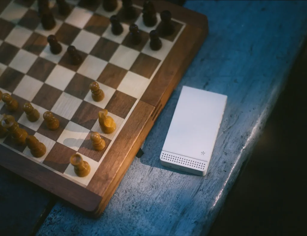 A white Light Phone rests beside a wooden chessboard on a weathered blue table, pairing the minimalist device with a quiet, analog set
