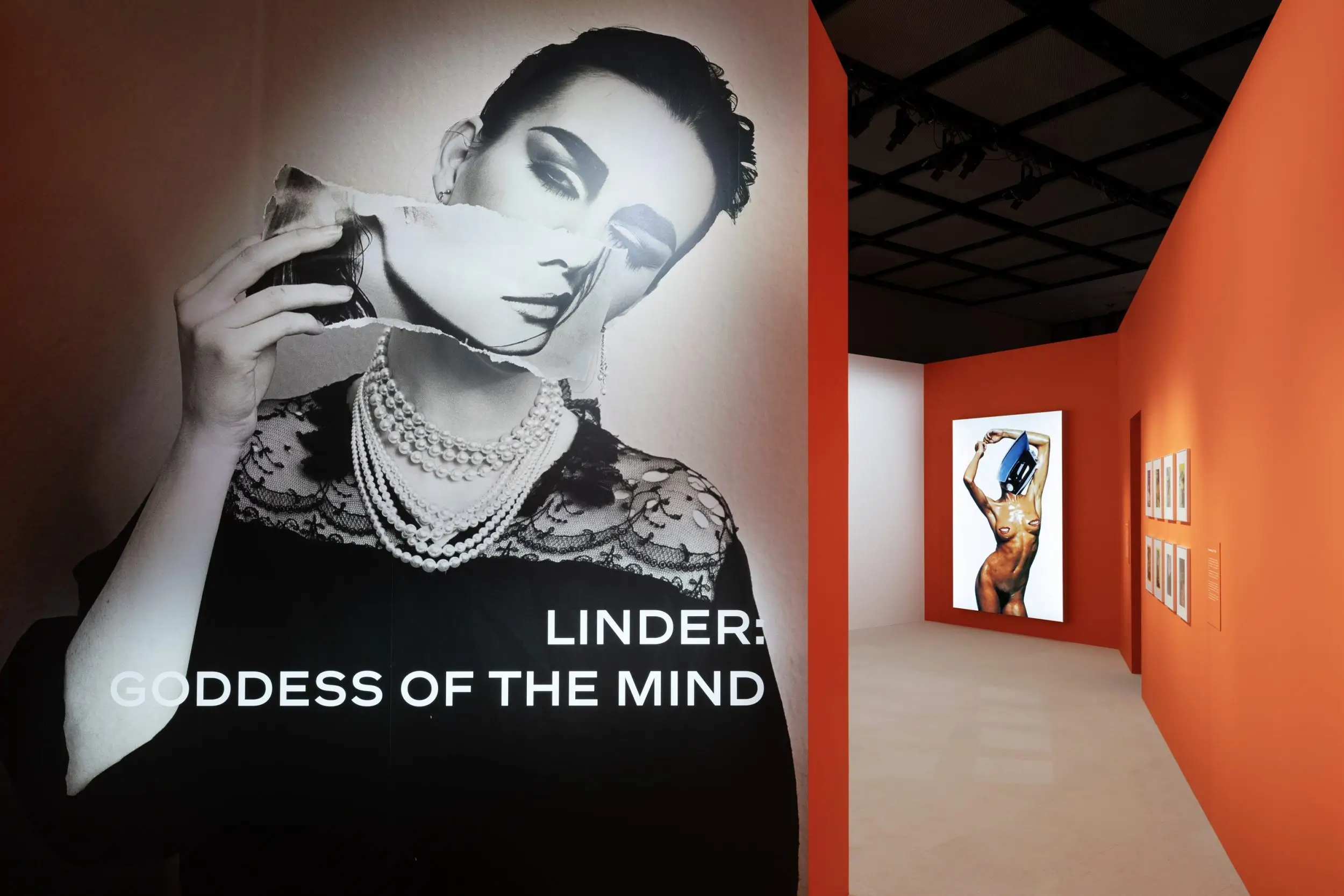 Entrance to the 'LINDER: GODDESS OF THE MIND' exhibition featuring a monumental black-and-white portrait with a torn collage effect, leading into a gallery with vivid orange walls and framed artworks.