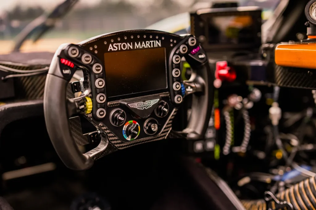 Close-up of an Aston Martin race car steering wheel featuring a built-in digital display, carbon-fiber construction, multifunction buttons, rotary switches, and performance controls designed for professional endurance racing