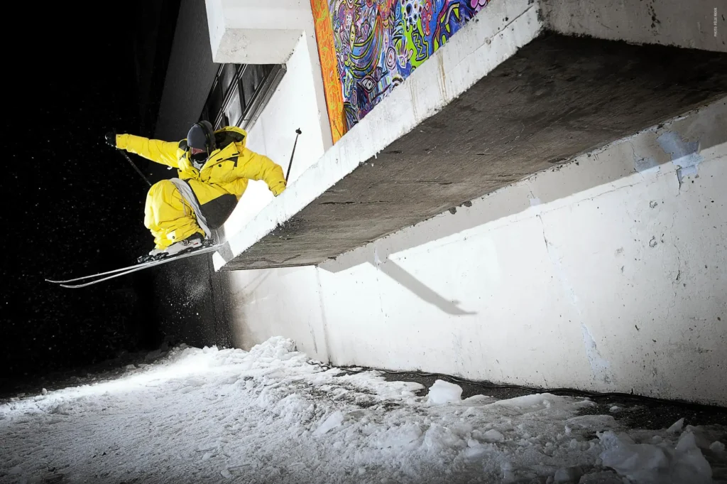 Nighttime urban skiing photograph of Martin Longhitano dropping from a concrete ledge beside a colorful mural, sending snow into the air during a technical street skiing session