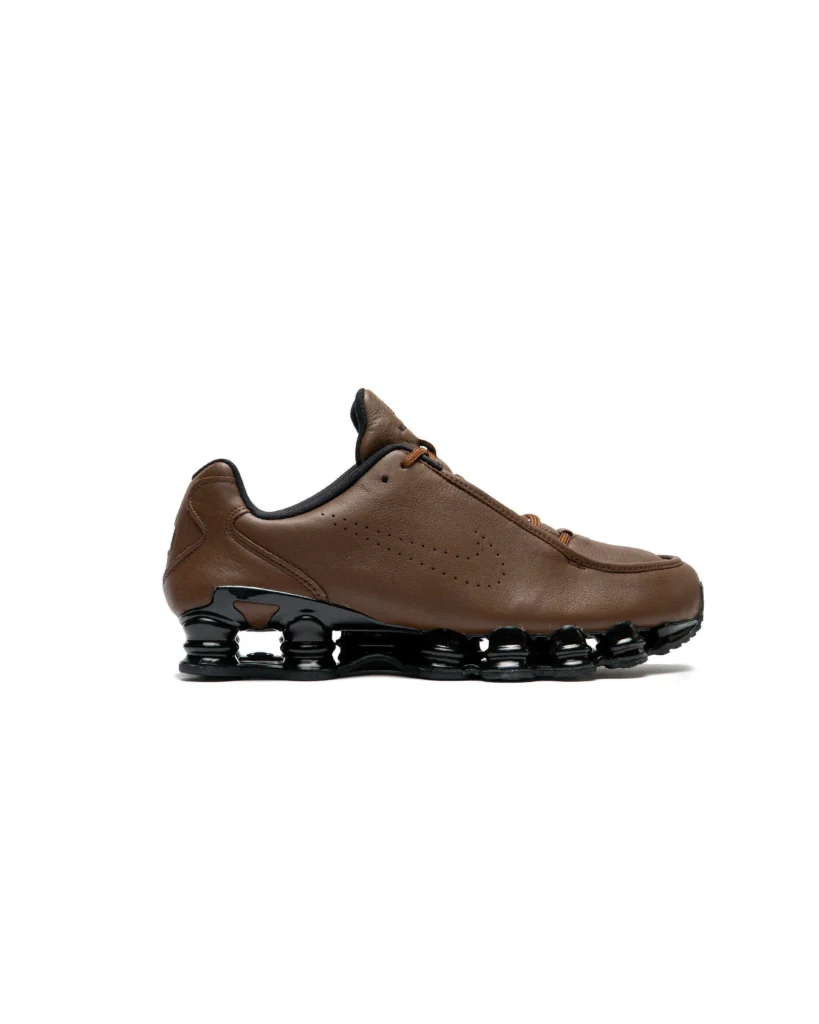 Side profile of the Nike Shox TL SE "Cacao Wow" (IR2097-200) featuring a premium brown leather upper with a perforated Swoosh, tonal laces, black collar lining, and glossy black full-length Shox cushion columns on a white background