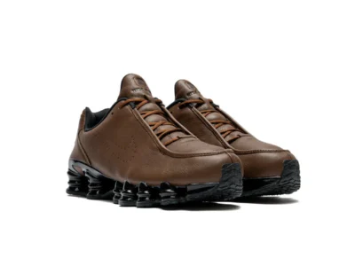 Front angled view of the Nike Shox TL SE "Cacao Wow" highlighting the moccasin-inspired toe shape, brown leather upper, tonal rope laces, embroidered Nike tongue branding, and glossy black full-length Shox cushio system