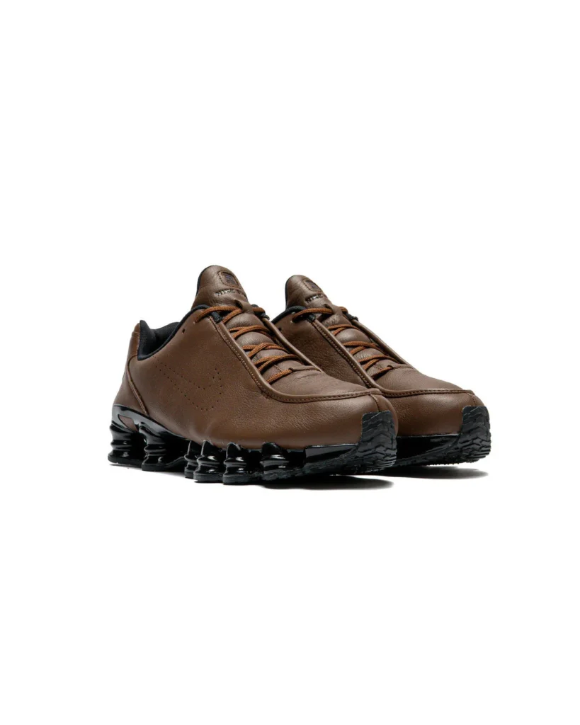 Front angled view of the Nike Shox TL SE "Cacao Wow" highlighting the moccasin-inspired toe shape, brown leather upper, tonal rope laces, embroidered Nike tongue branding, and glossy black full-length Shox cushio system