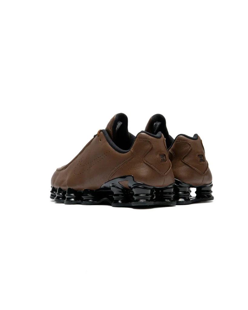 Rear three-quarter view of the Nike Shox TL SE "Cacao Wow" displaying the padded heel construction, embossed Nike heel branding, brown leather upper, and signature glossy black full-length Shox cushion platform on a white background