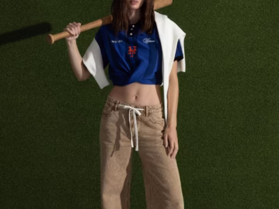 A model stands against a textured artificial turf backdrop holding a wooden baseball bat over one shoulder. She wears a cropped blue New York Mets–inspired jersey with orange logo detailing, wide-leg tan drawstring jeans, and a white sweater draped over her shoulders, blending baseball aesthetics with contemporary streetwear style
