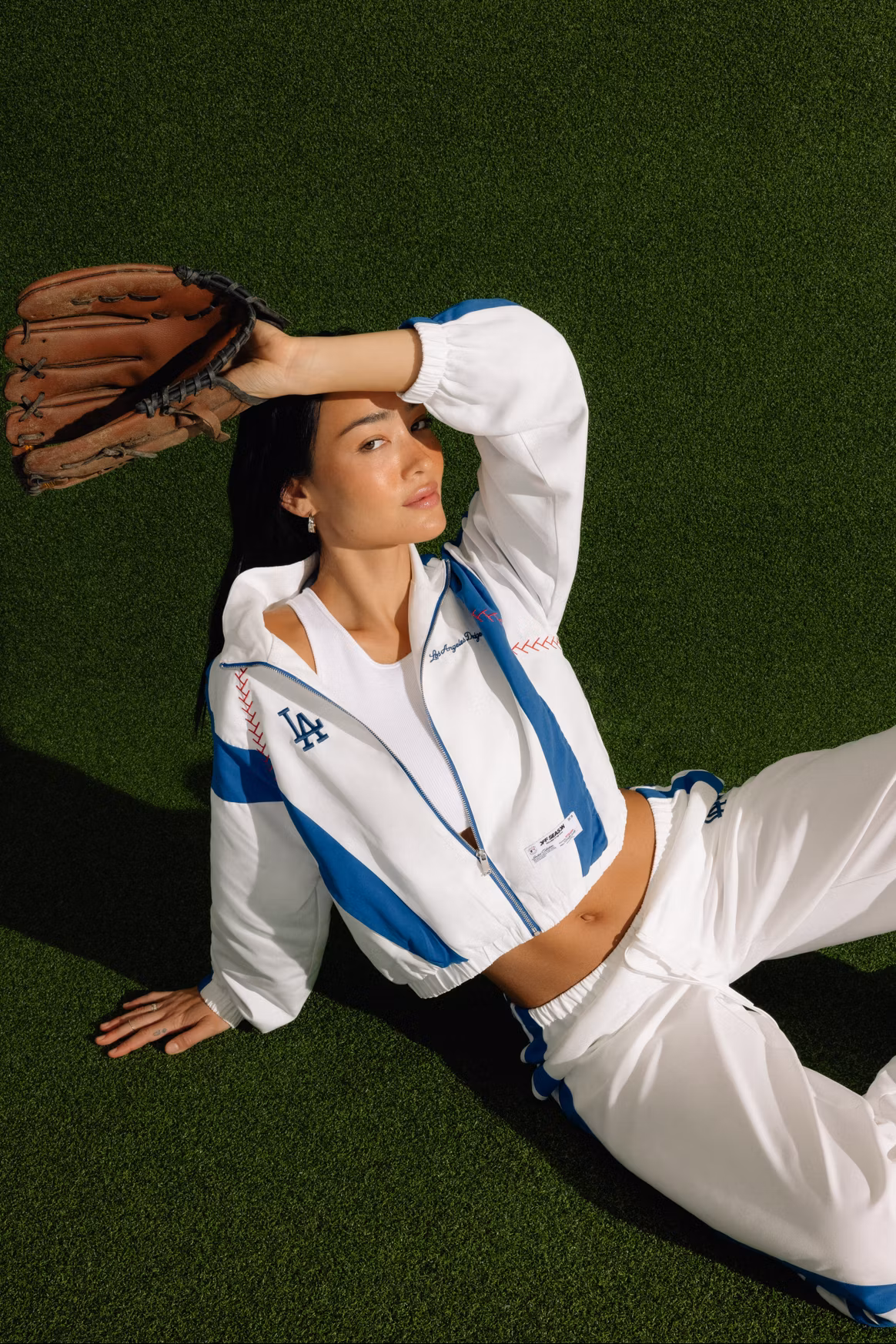Model reclining on artificial turf wearing a white and blue Los Angeles Dodgers-inspired tracksuit with a cropped tank, holding a worn leather baseball glove over her forehead in a fashion campaign blending athletic heritage with contemporary streetwear.