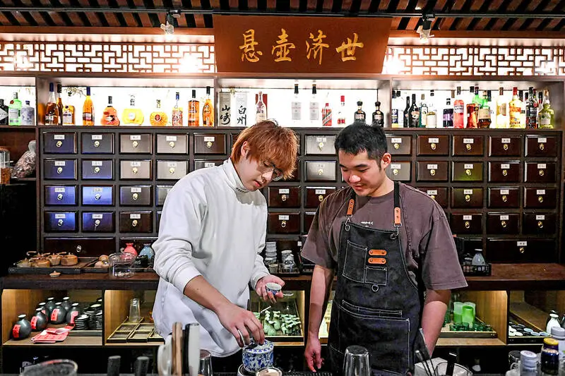 Two staff members prepare herbal ingredients behind a traditional Chinese medicine-inspired bar, with rows of wooden apothecary drawers, shelves of spirits, tea ware, and Chinese calligraphy signage creating a blend of wellness clinic and cocktail bar aesthetics.