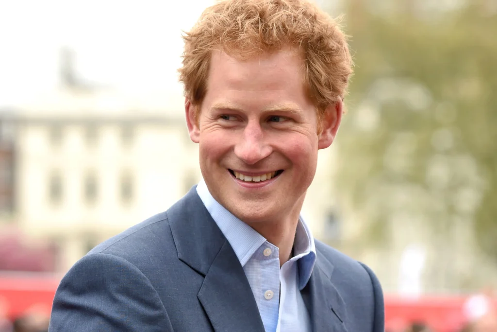 Prince Harry with short ginger hair looks over his shoulder while wearing a navy blue suit jacket and a light blue dress shirt. The softly blurred outdoor background features historic architecture and trees, drawing attention to his relaxed expression and polished appearance
