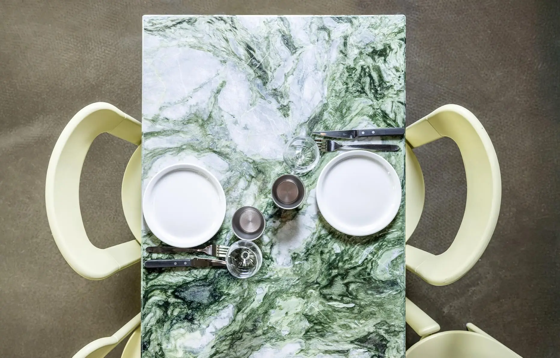 Overhead view of a square green marble café table set for two with white plates, glassware, cutlery, and pale green chairs arranged in a clean, minimalist dining setting.