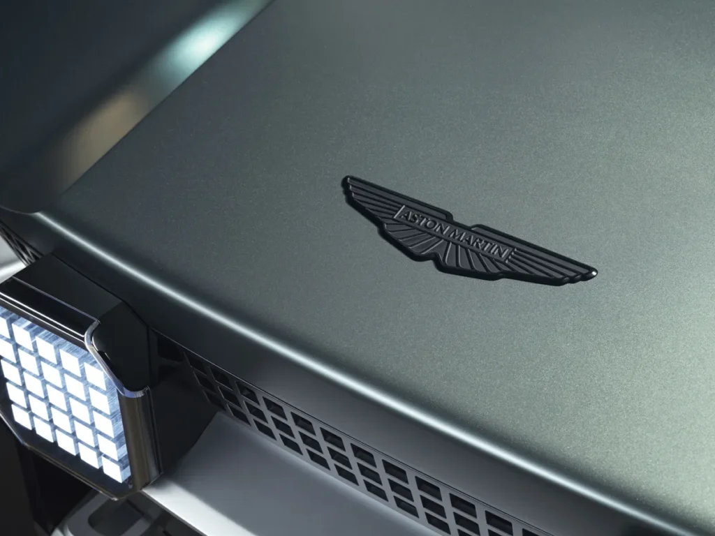 Detail of the front hood displaying Aston Martin's black winged emblem on matte metallic green paint, alongside a square LED auxiliary light module and rugged grille vents that reinforce the concept's off-road style
