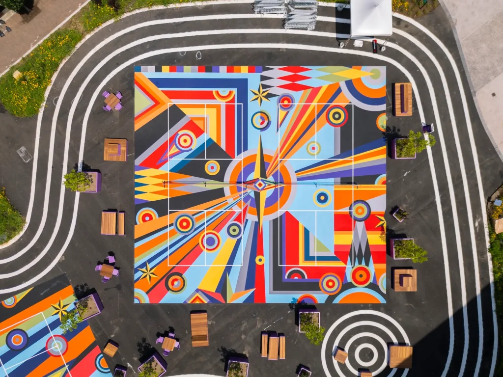 Top-down aerial view of a vibrant square sports court mural composed of bold geometric shapes, concentric circles, and radiating orange, blue, yellow, red, and gray patterns, surrounded by benches, planters, and looping white pathways