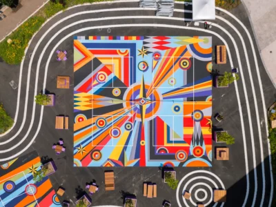 Top-down aerial view of a vibrant square sports court mural composed of bold geometric shapes, concentric circles, and radiating orange, blue, yellow, red, and gray patterns, surrounded by benches, planters, and looping white pathways