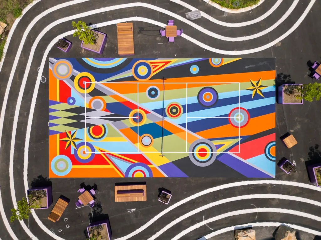 Overhead view of a rectangular outdoor sports court painted with intersecting colorful stripes, concentric circles, and star motifs, framed by curved white pathway lines, benches, purple planters, and landscaped seating areas